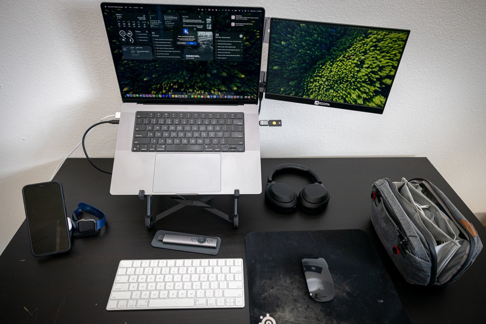 A MacBook on a Roost stand beside a portable second monitor, with a Magic Keyboard, mouse, headphones and a Peak Design pouch on a black desk